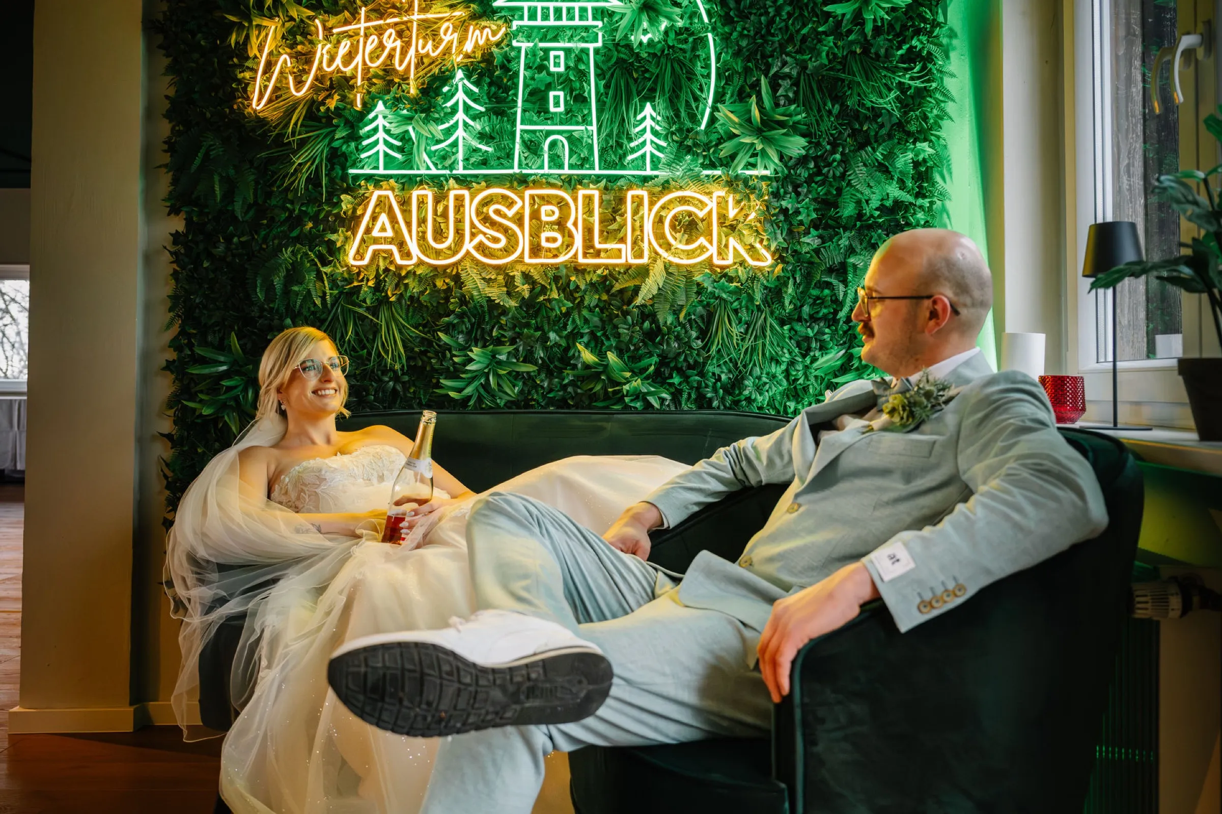 Brautpaar genießt einen entspannten Moment vor der goldenen Neon-Mooswand